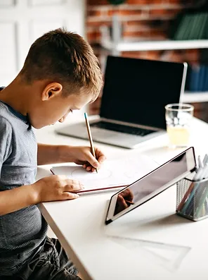 Menino estudando, escrevendo em um caderno sobre a mesa com notebook e tablet.