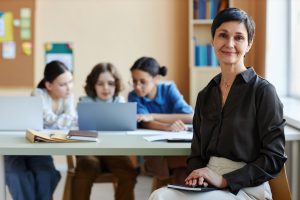 Professora sentada em sala de aula com alunos ao fundo