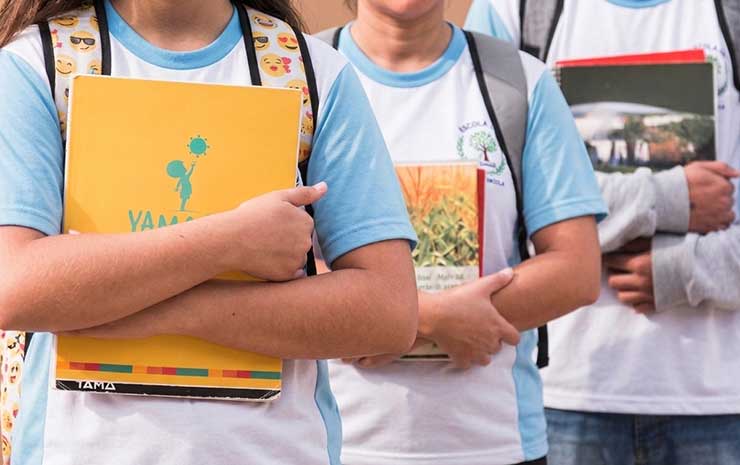 Estudantes da educação básica, com uniforme azul-claro, seguram livros e cadernos em ambiente escolar externo, representando a realidade da rede pública e o contexto do Censo Escolar 2025.